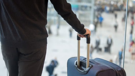 Young handsome man in 20s waiting for flight, standing in modern airport terminal with crowd on background, holding handle of luggage bag, wearing casual clothes, rear view, close up