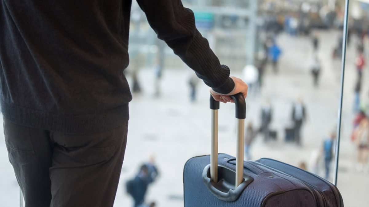 Young handsome man in 20s waiting for flight, standing in modern airport terminal with crowd on background, holding handle of luggage bag, wearing casual clothes, rear view, close up