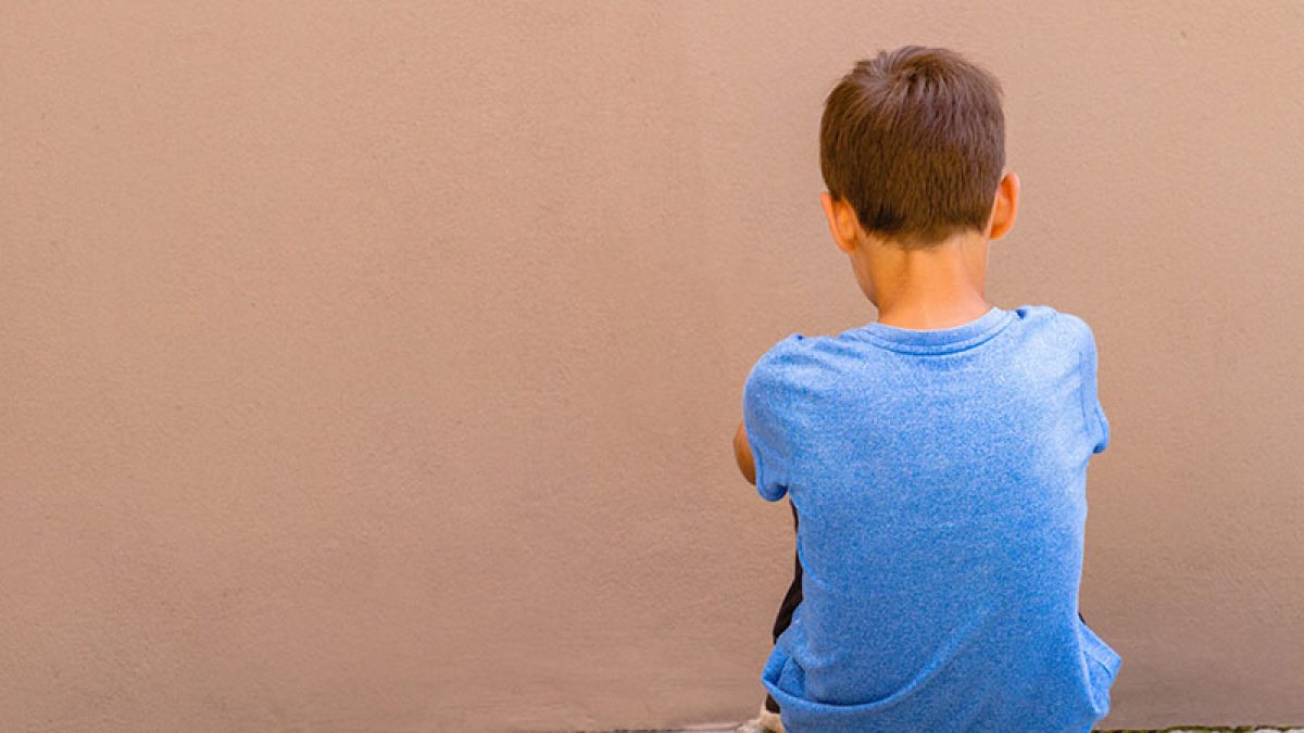 Sad alone boy sitting on the ground behind the wall outdoor.